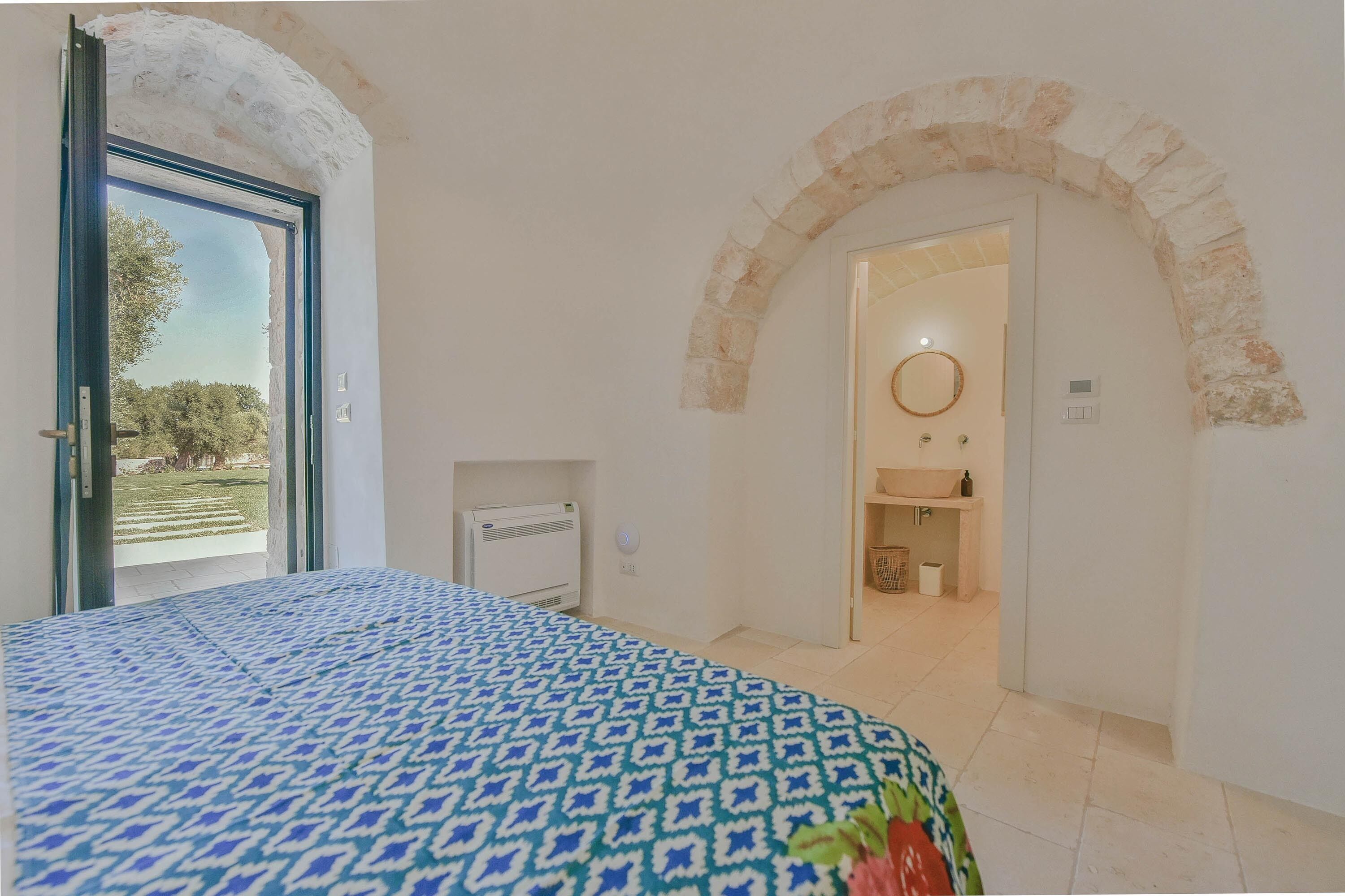 Un dormitorio con una colcha estampada, un arco de piedra, una vista por una puerta y un baño con lavamanos y espejo.