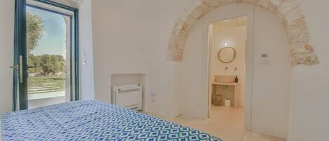 Un dormitorio con una colcha estampada, un arco de piedra, una vista por una puerta y un baño con lavamanos y espejo.