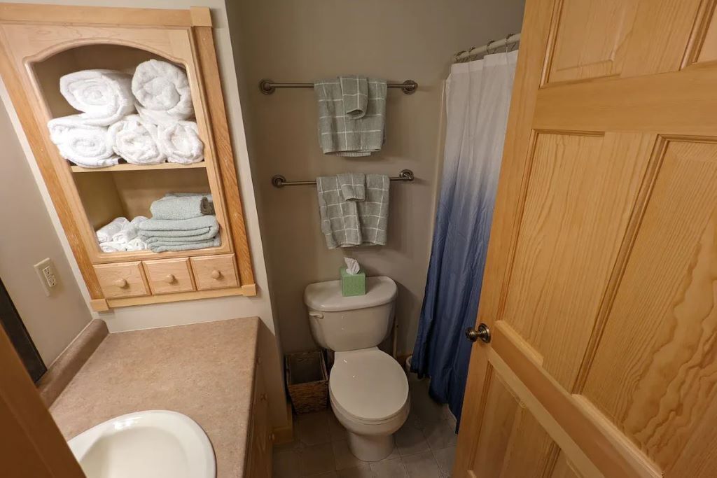 A small bathroom with a toilet, a towel rack with folded towels, a wooden cabinet with more towels, and a blue shower curtain.
