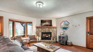 A living room with a stone fireplace, a large window with blinds, a couch, a coffee table, and a bookshelf. - Gatlinburg Mountain View Cabin | Hot Tub & Arcade (Gatlinburg)