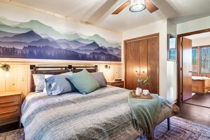 Ein Schlafzimmer mit einem Bett, einem Holzkopfteil, Nachttischen, Wandmalerei, Deckenventilator und einer Tür zu einem anderen Raum.