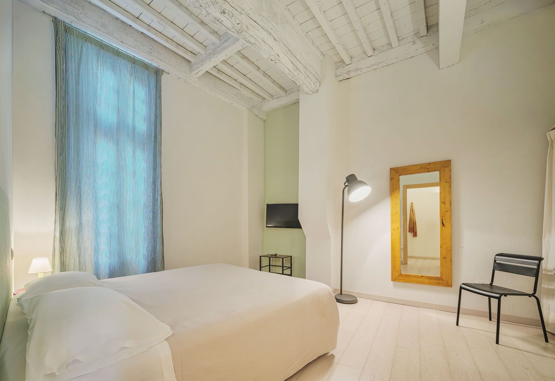 A hotel room with a bed, a desk, a chair, a lamp, and a mirror.
