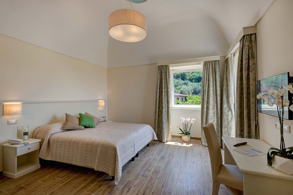 Un dormitorio con cama, mesita de noche, lámpara, silla, escritorio y ventana con cortinas.