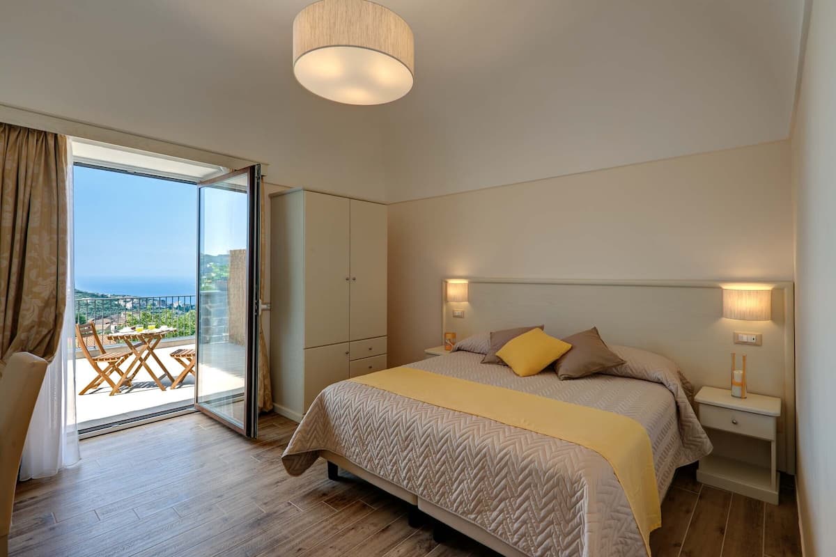 Un dormitorio con una cama grande, una mesita de noche, una lámpara, un armario y un ventanal corredero que da a un balcón con vistas.