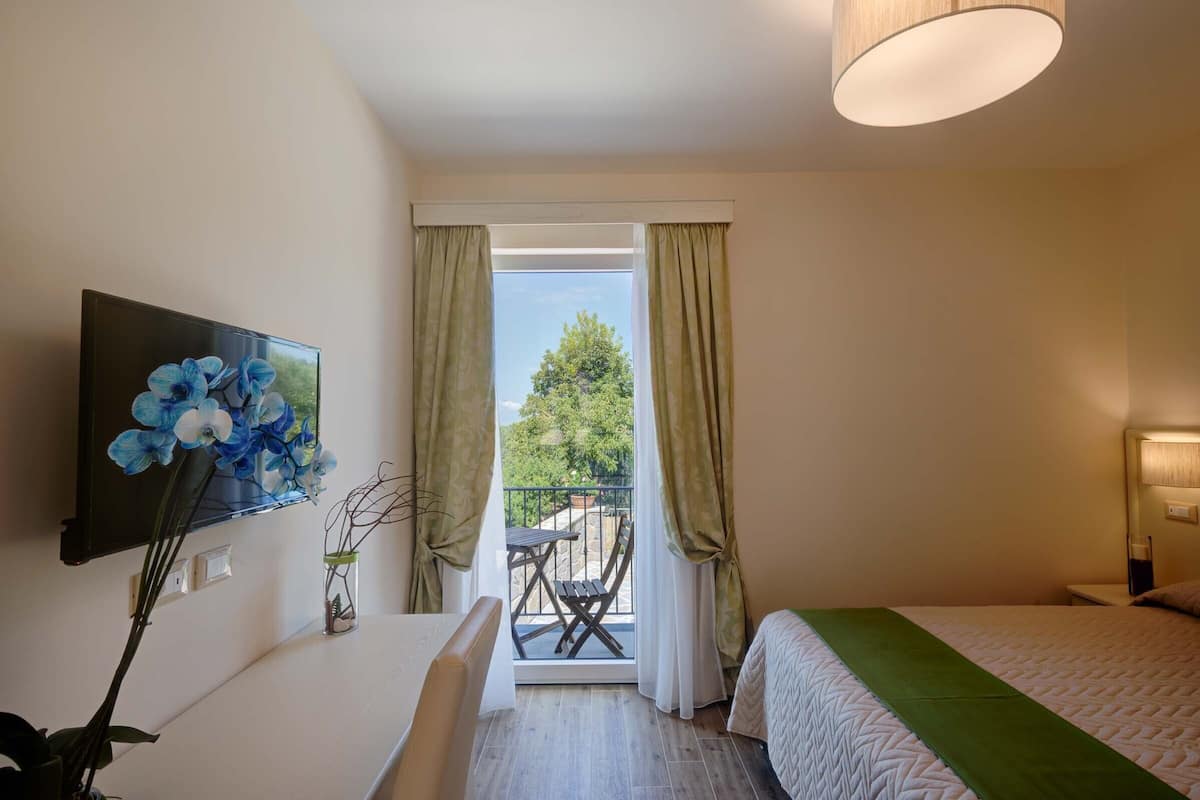 Habitación de hotel con cama, escritorio, televisión, balcón con mesa y sillas, y vista a árboles.