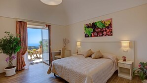 Een slaapkamer met een bed, een bureau, een stoel, een kamerplant en een schilderij van druiven aan de muur.