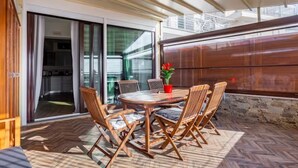 Een terras buiten met een houten tafel en stoelen, een bloempot en uitzicht op een gebouw door glazen deuren.