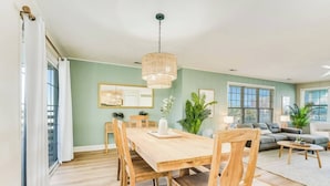 A dining area with a wooden table and chairs, a hanging light fixture, and a view of the living room with a sofa and plants. - Ocean & Sound Views, Game Room, Steps to Beach! (Surf City)