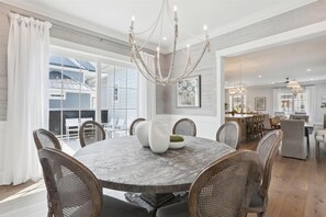 A dining area with a round table, six wicker chairs, a chandelier, and large windows.
