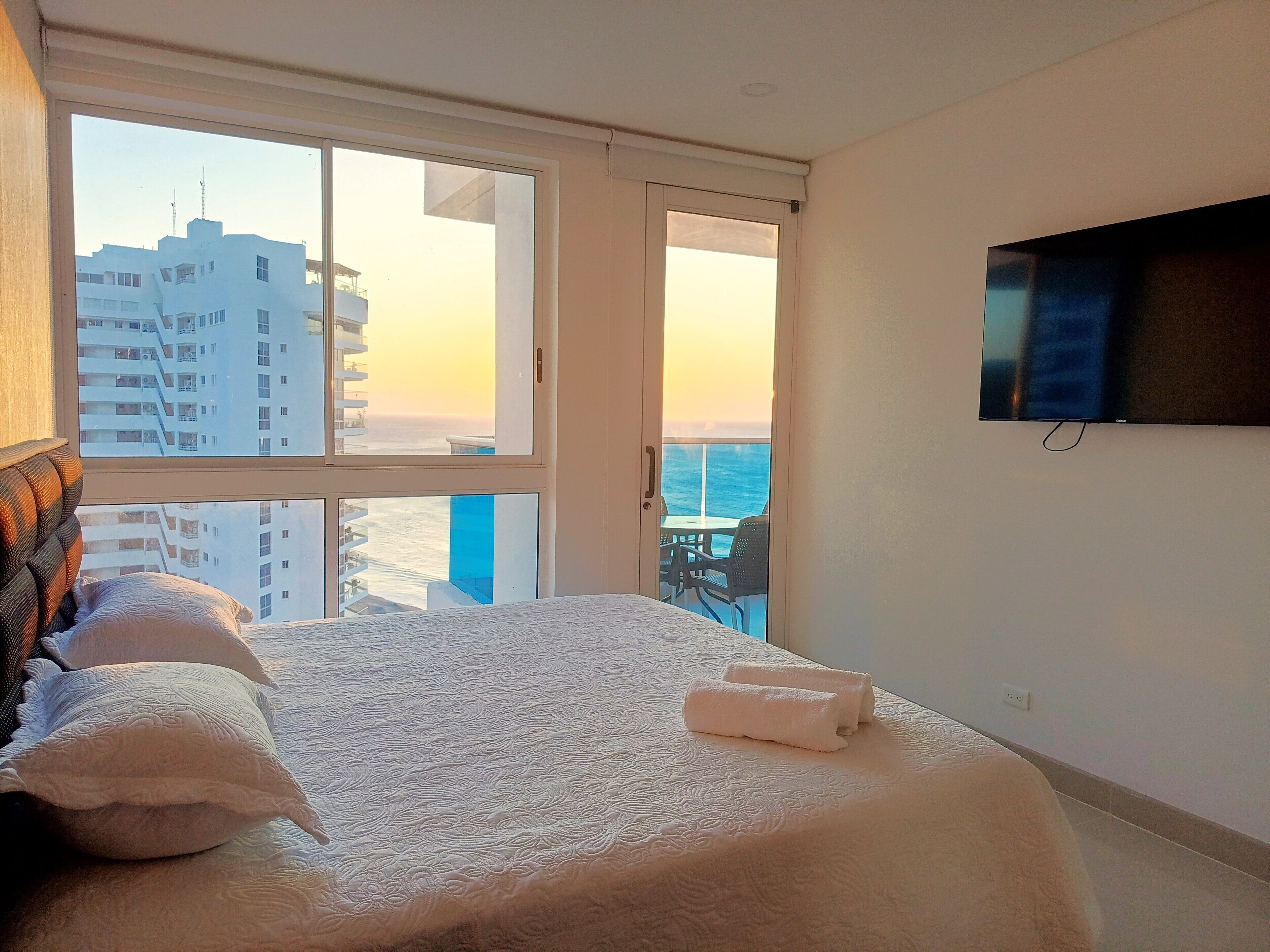 A hotel room with a bed, pillows, a TV, and a balcony with a view of the city and ocean.