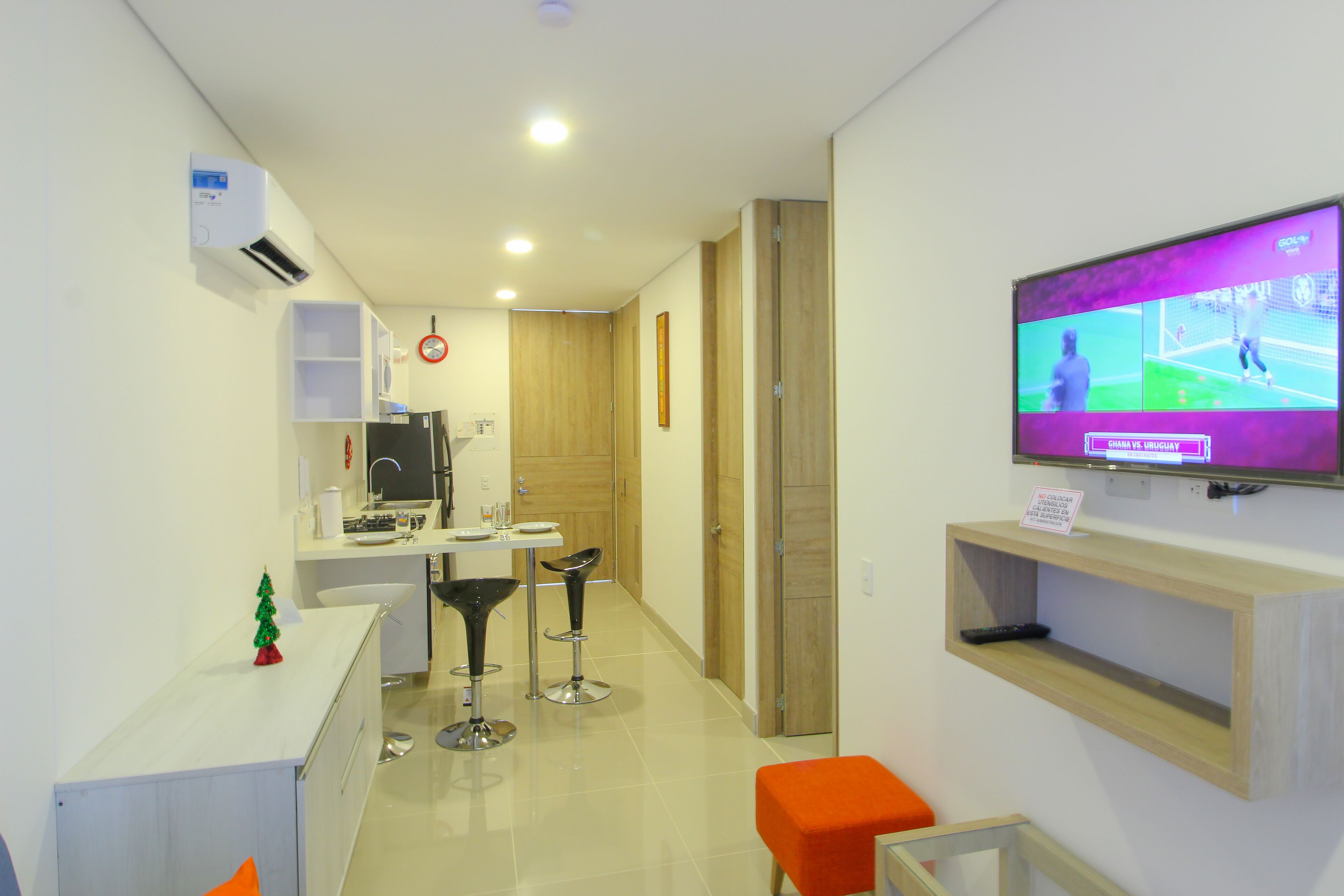 A modern hotel room with a TV displaying a sports game, a small kitchen area, and a seating area with a bar stool.