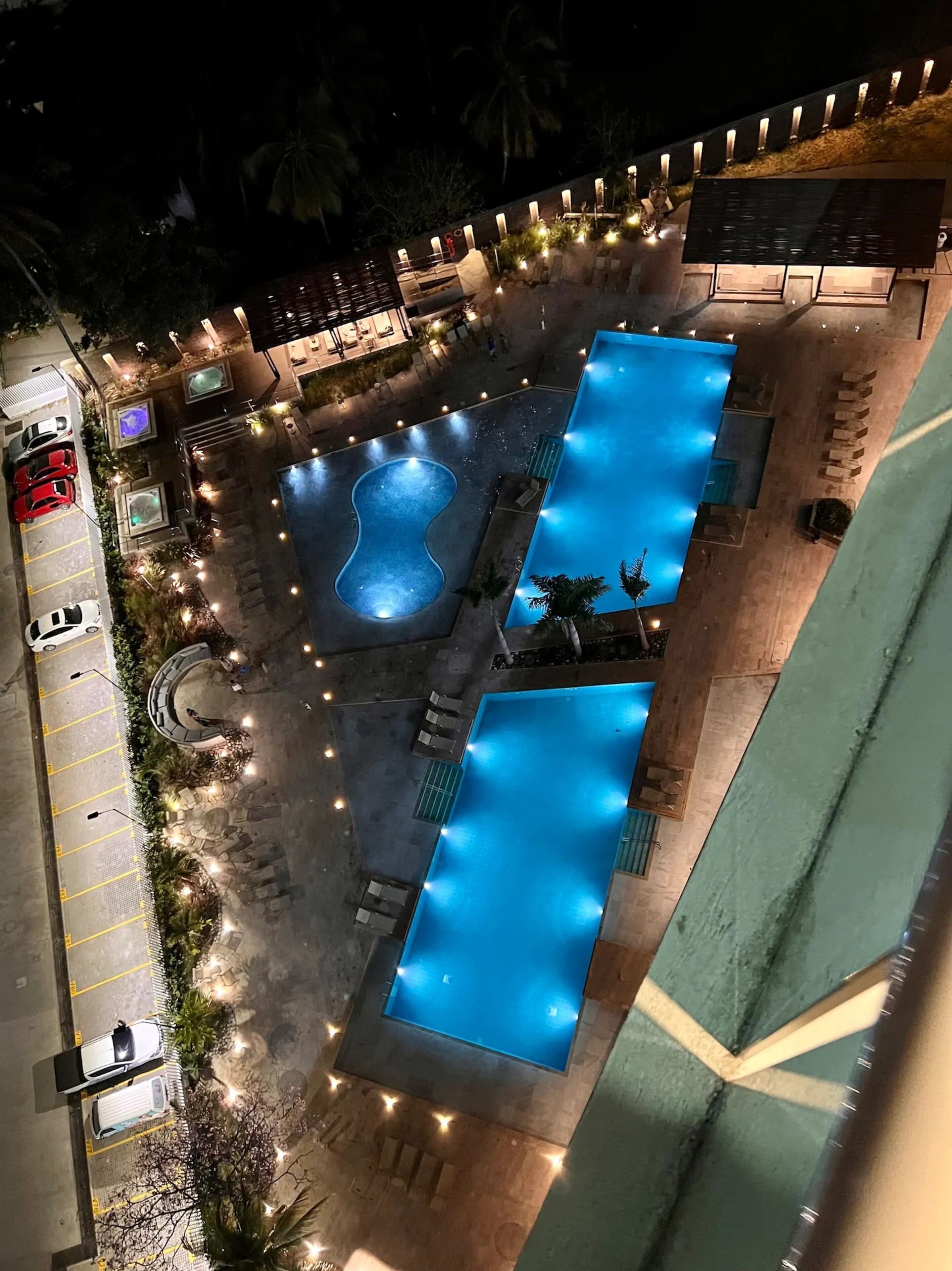 An aerial view of a hotel with multiple swimming pools, a parking area, and a landscaped garden.