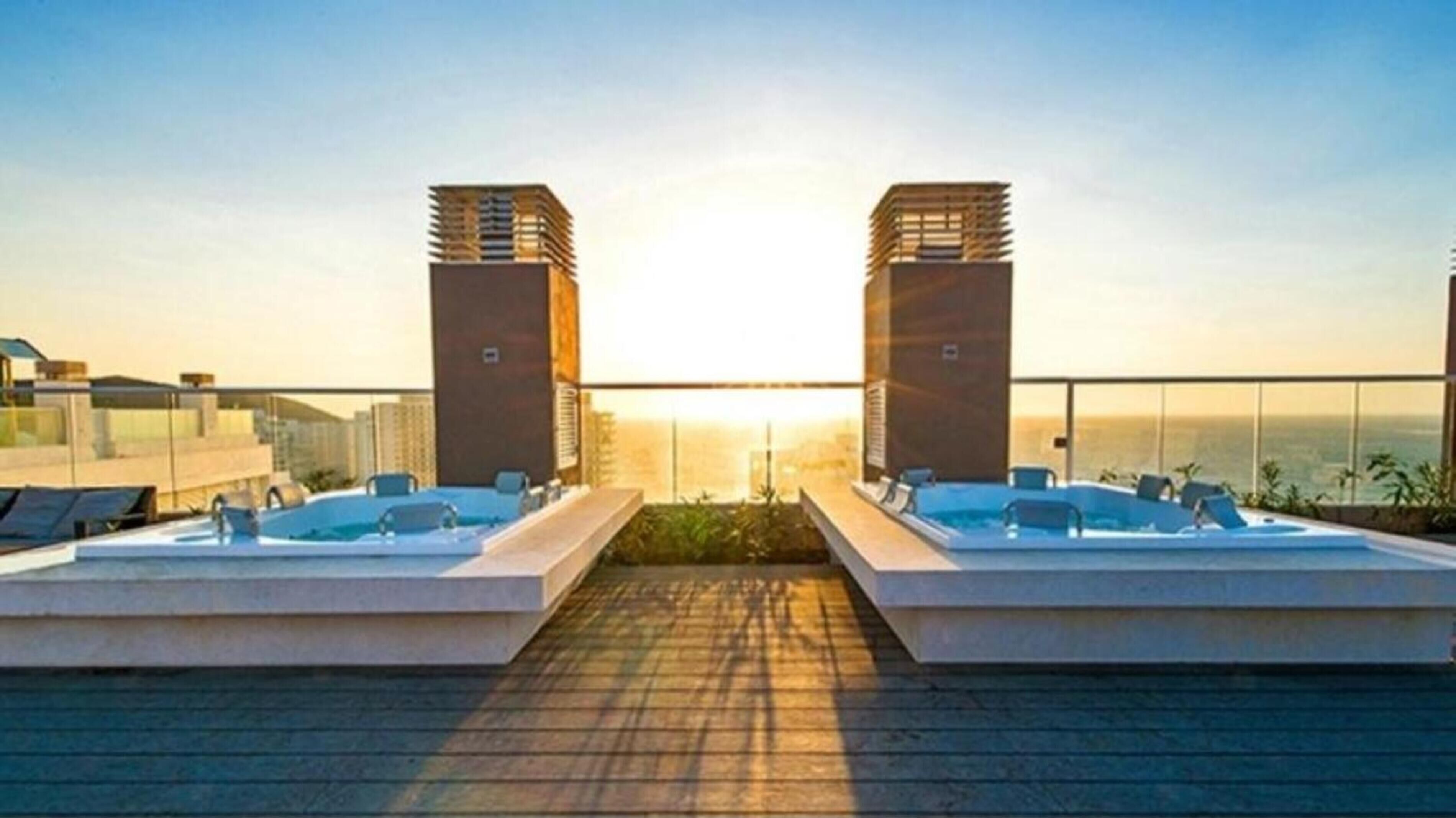 Un área de piscina en la azotea con dos jacuzzis, cielo despejado y un atardecer de fondo.