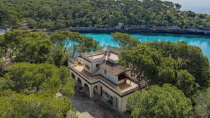 Een villa in mediterrane stijl met een terracotta dak, stenen muren en een stenen binnenplaats. Het heeft een balkon met buitenmeubilair en uitzicht op een turquoise zee en weelderige begroeiing.