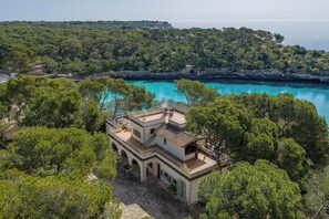 Une villa de style méditerranéen, dotée d’un toit en tuiles de terre cuite, de murs en pierre et d’une cour intérieure en pierre. Elle possède un balcon avec des sièges d’extérieur et offre une vue sur une mer turquoise et une végétation luxuriante.