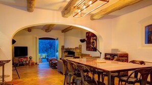 Uma ampla área de refeições com uma mesa comprida e cadeiras, uma lareira e vista para uma área externa através de um arco.