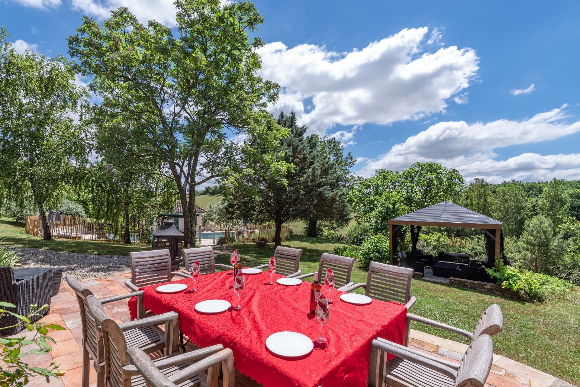 An outdoor dining area with a red tablecloth, wooden chairs, and a gazebo in the background.
