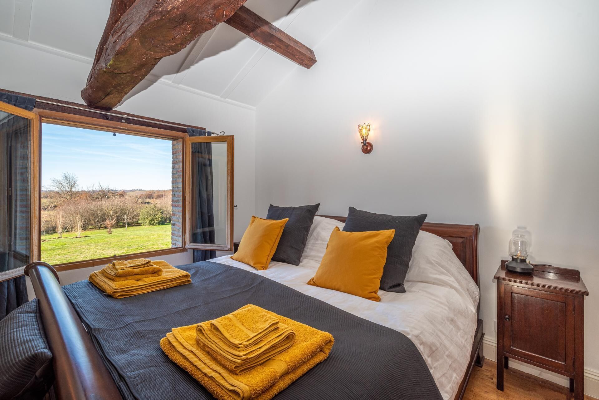 Un dormitorio con una cama grande, cabecera de madera, mesita de noche y vistas a un paisaje verde a través de una ventana abierta.