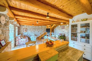 A rustic interior with a stone wall, wooden ceiling, a dining area with a bench, a kitchen with cabinets, and a staircase.