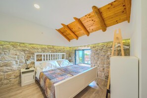 Une chambre avec un mur en pierre, un plafond en bois, un lit recouvert d’une couverture à motifs, une table de chevet et une armoire.