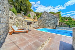 A stone villa with a swimming pool, two lounge chairs, and a small table with an umbrella.