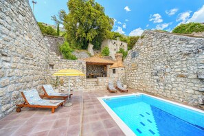 Un espace piscine entouré de murs en pierre, avec des chaises longues et un petit coin salon abrité.