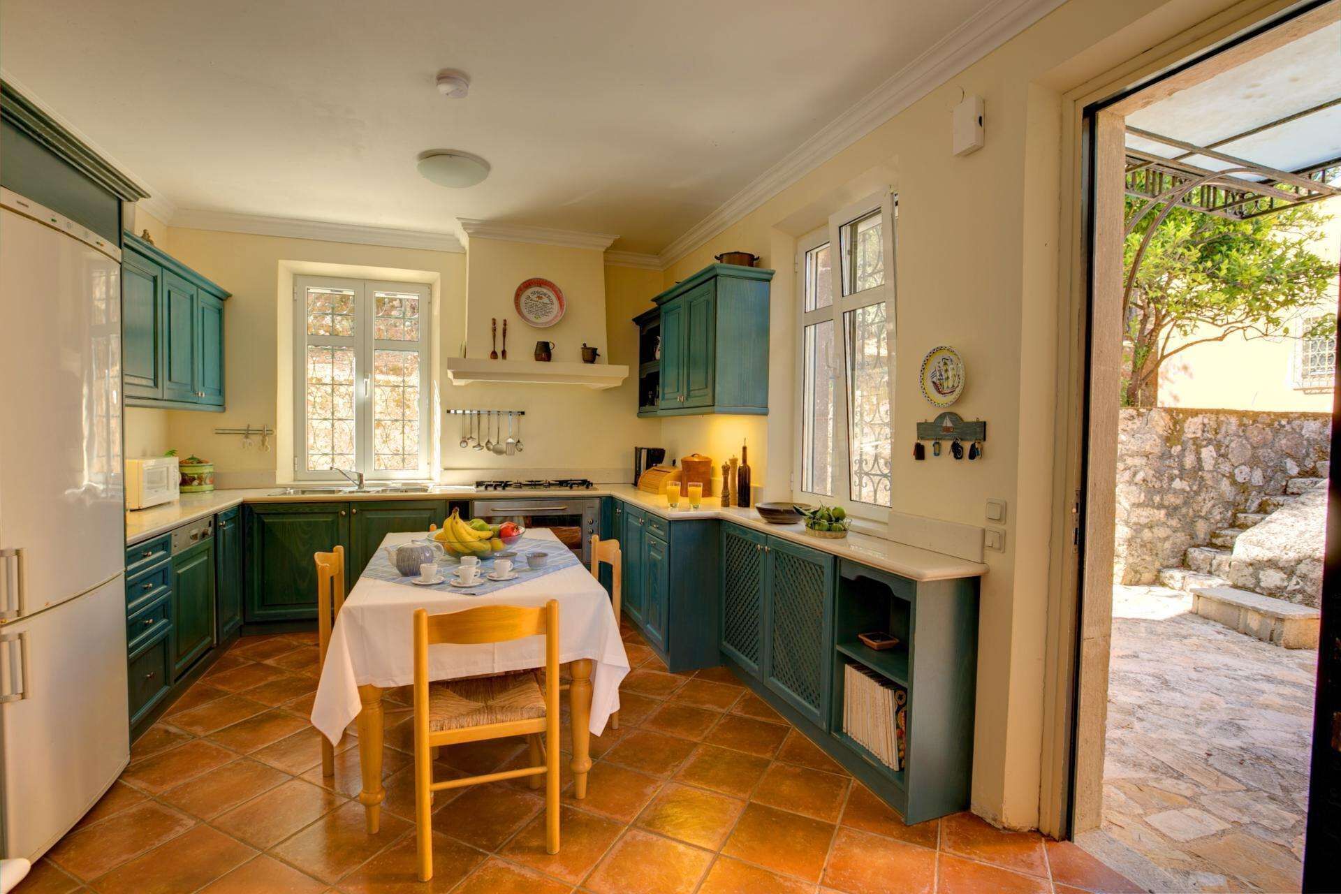 Une cuisine avec des meubles de couleur turquoise, une table à manger recouverte d’une nappe blanche et une vue sur un mur en pierre à l’extérieur.