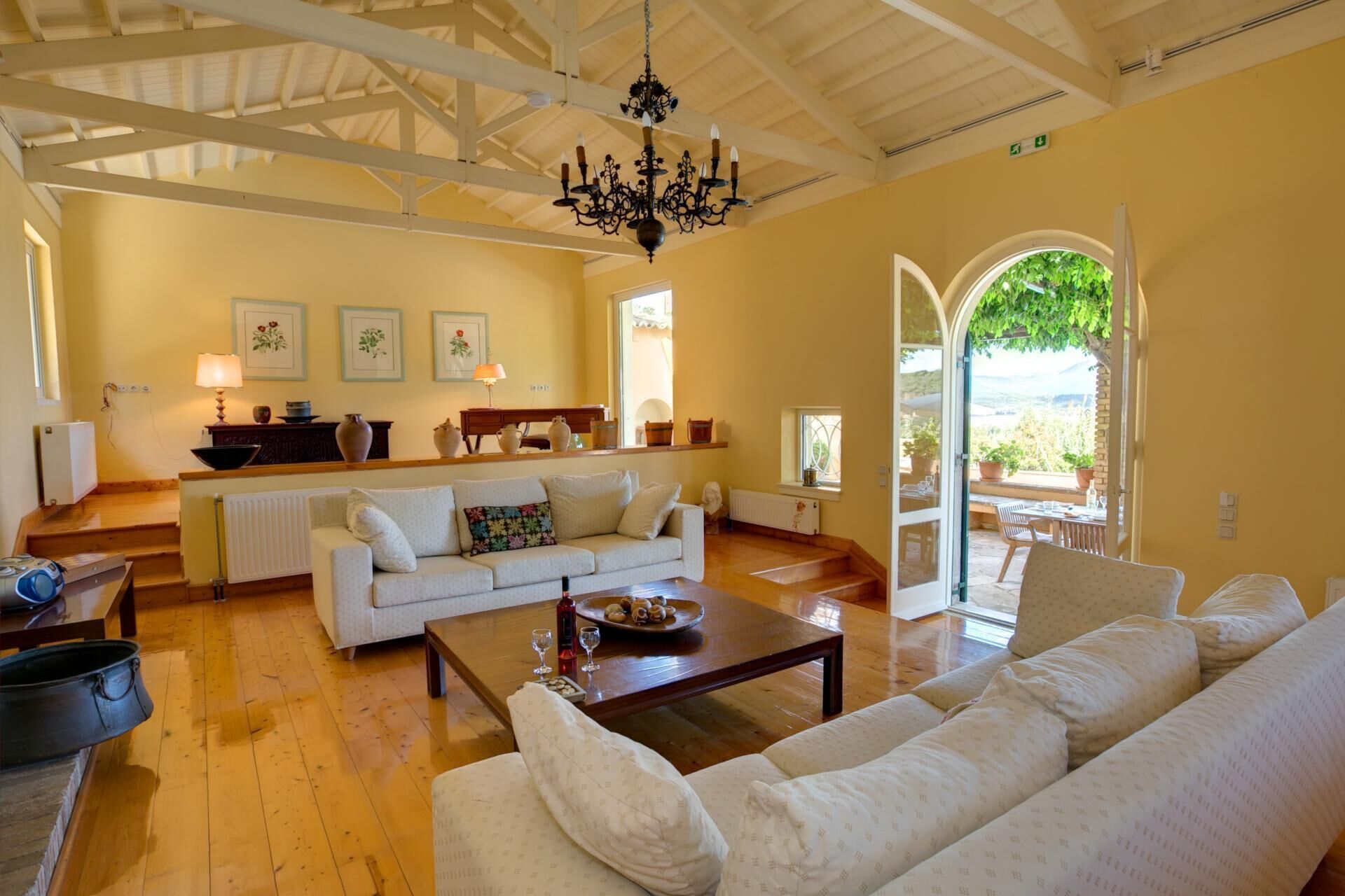 Amplia sala de estar con un sofá blanco, una mesa de centro de madera y un candelabro. Hay una zona de cocina con mostrador y diversos utensilios. Un ventanal ofrece vistas al exterior.