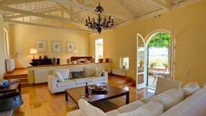 Amplia sala de estar con un sofá blanco, una mesa de centro de madera y un candelabro. Hay una zona de cocina con mostrador y diversos utensilios. Un ventanal ofrece vistas al exterior.