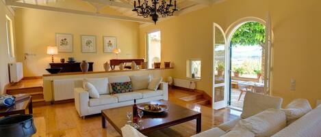 Amplia sala de estar con un sofá blanco, una mesa de centro de madera y un candelabro. Hay una zona de cocina con mostrador y diversos utensilios. Un ventanal ofrece vistas al exterior.
