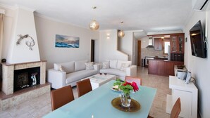 A spacious living area with a sofa, a dining table with chairs, a fireplace, and a kitchen in the background. - Pegasus - Three Bedroom Villa, Sleeps 6 (Spartochori)