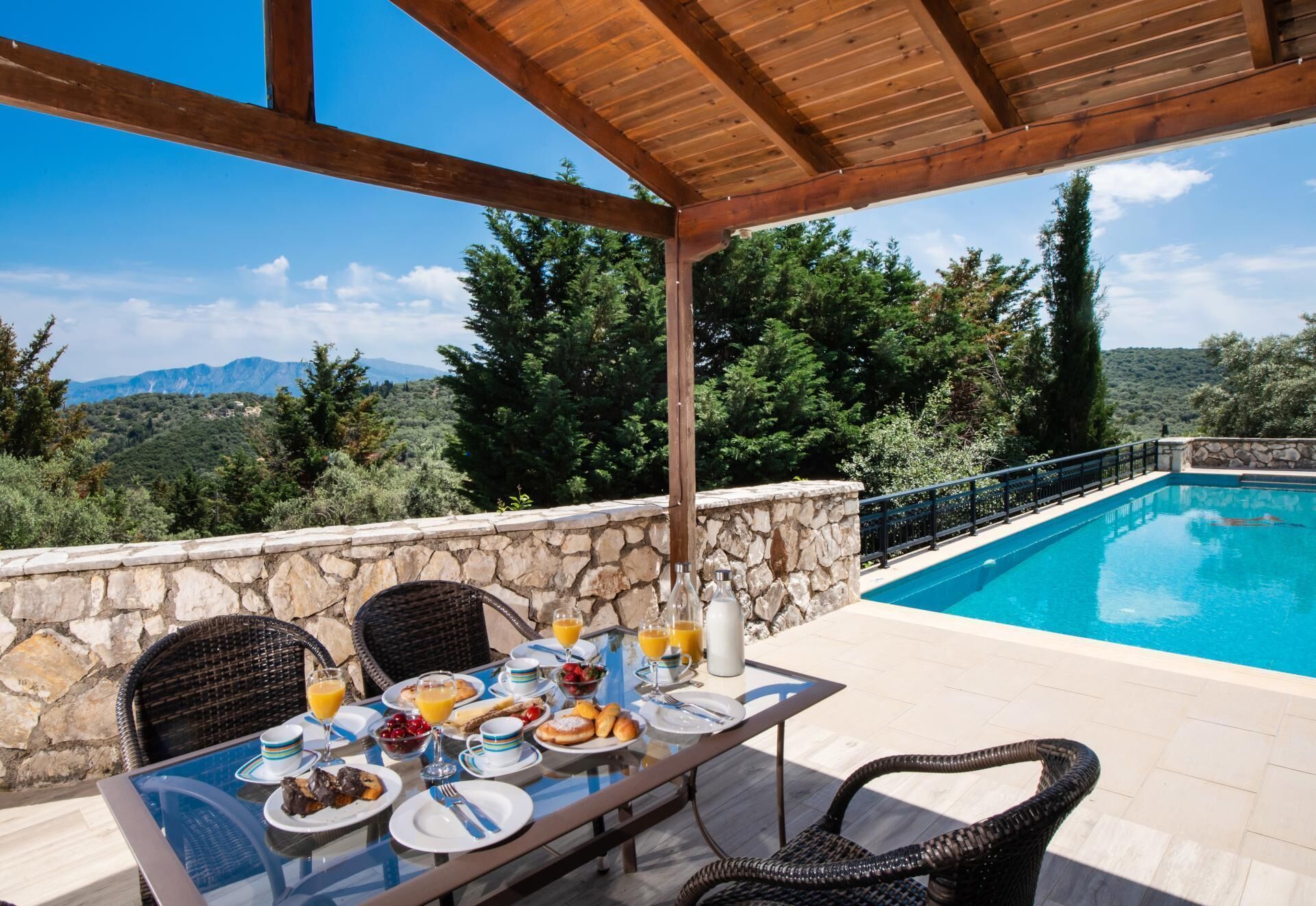 A dining table with breakfast items, a stone wall, and a poolside view of a mountainous landscape.