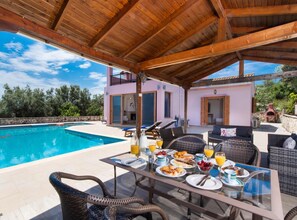 A covered outdoor dining area with a pool, wicker furniture, and a table set for breakfast.