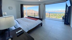 Una habitación de hotel moderna con una cama grande, una mesita de noche con lámpara, un control remoto y vistas a un paisaje montañoso desde un balcón.