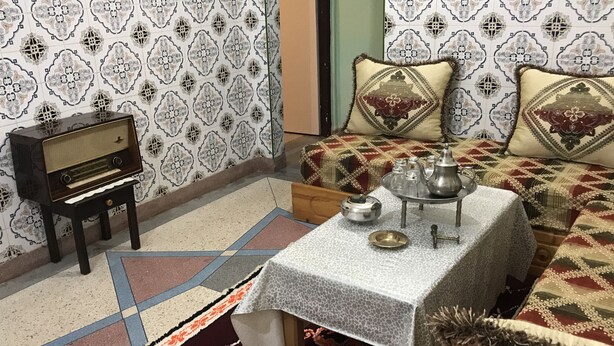 Una habitación con papel tapiz estampado, una radio antigua, una mesa con tetera y vasos, y una cama con almohadas decorativas.