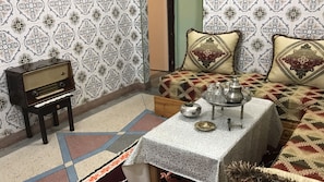 Una habitación con papel tapiz estampado, una radio antigua, una mesa con tetera y vasos, y una cama con almohadas decorativas.