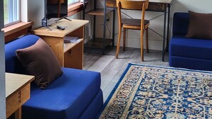 A cozy living room with a blue sofa, a wooden desk, and a computer. There are large windows with blinds, a rug, and a painting on the wall. - Lake Champlain Tiny House Paradise Private & Beautiful! (Alburgh)