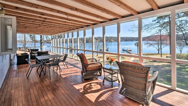 A covered deck with wooden beams, wicker furniture, and large windows overlooking a lake. - Stunning Seneca Home w/ Lake Keowee Access! (Seneca)