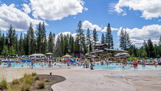 A large outdoor swimming pool surrounded by trees, with a water slide and lounge areas. - Walk to the Village, Hot Tub, SHARC pass, AC, Games, Bikes, Grill, Pet-Friendly (Sunriver)