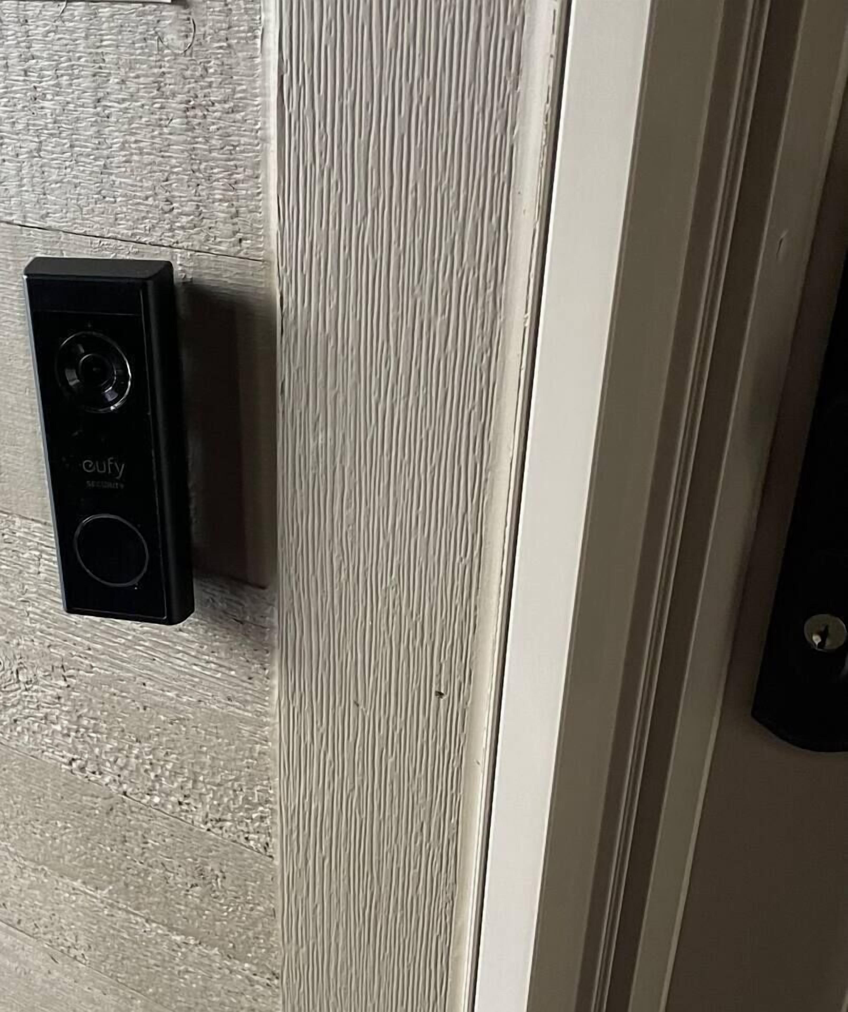 Puerta con pared blanca texturizada y un sistema de intercomunicación negro a la izquierda.