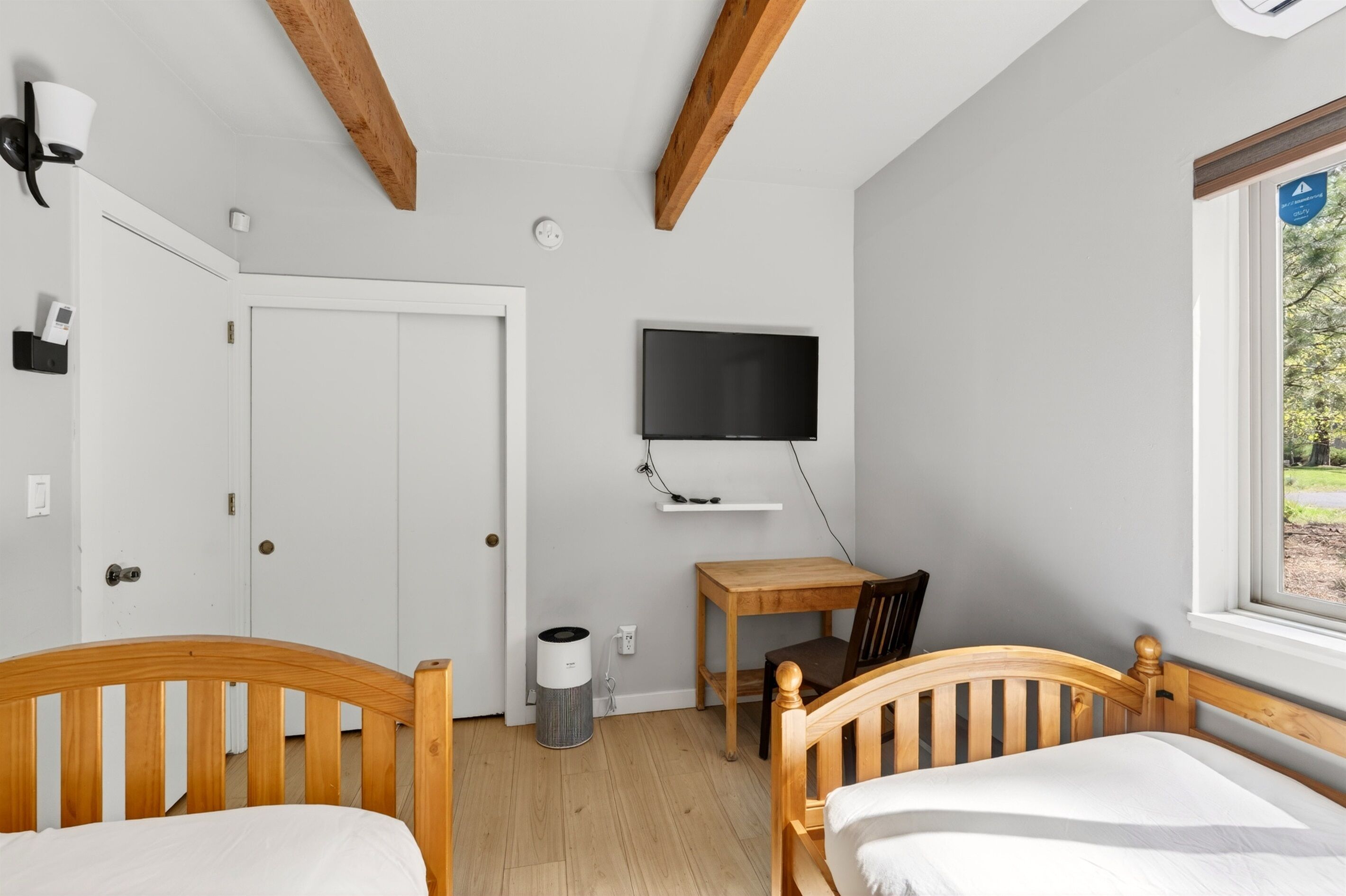 A bedroom with two wooden beds, a TV mounted on the wall, a small wooden table, and a window with a view of greenery.