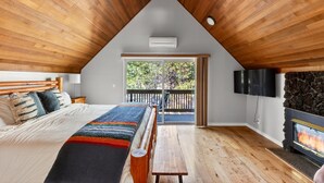 A bedroom with a sloped ceiling, a bed with a colorful blanket, a bench, a fireplace, and a TV mounted on the wall. - Walk to the Village, Hot Tub, SHARC pass, AC, Games, Bikes, Grill, Pet-Friendly (Sunriver)