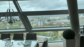 A modern dining area with a glass table and chairs, a hanging light fixture, and a view of a cityscape through large windows. - Charming 2-bed Apartment in London (London)