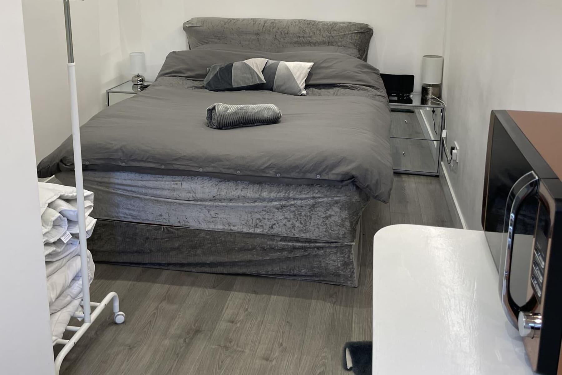 Una cama bien hecha con ropa de cama gris, una mesita de noche con lámpara y un escritorio pequeño con ordenador.