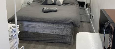 Una cama bien hecha con ropa de cama gris, una mesita de noche con lámpara y un escritorio pequeño con ordenador.