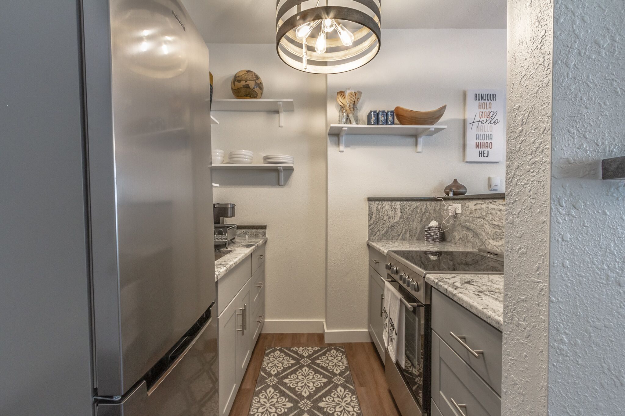 Una cocina compacta con un refrigerador de acero inoxidable, horno empotrado y encimera de granito. Hay armarios blancos y una alfombra estampada en el suelo.