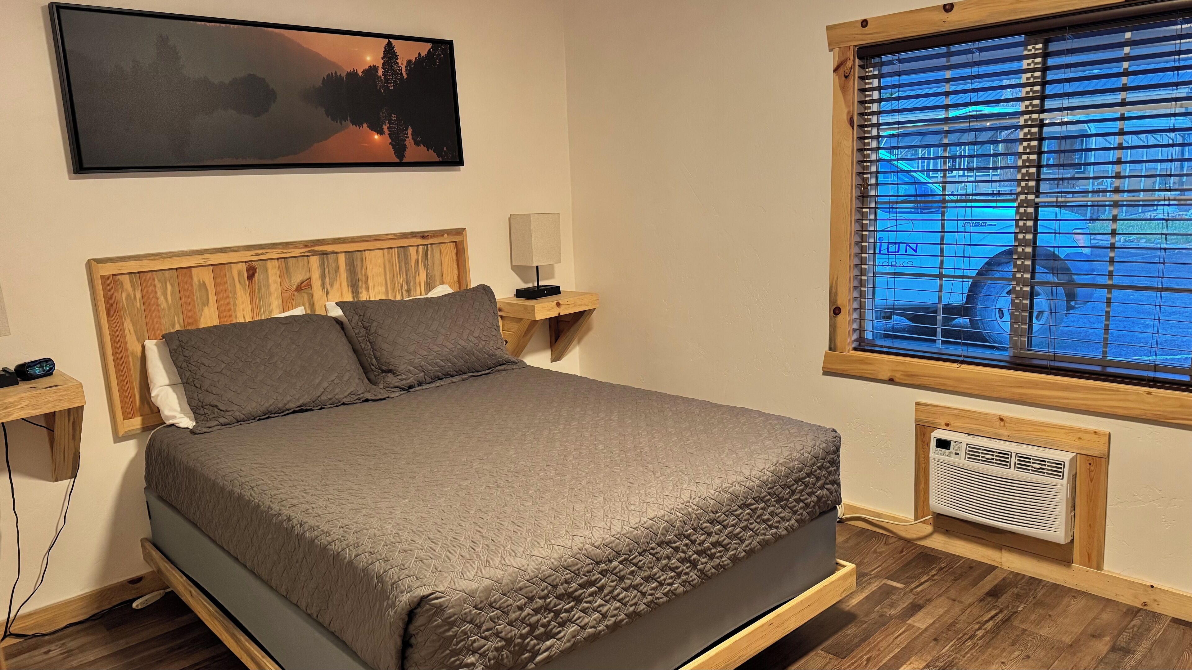 A bedroom with a wooden headboard, a bed with a quilted cover, a nightstand with a lamp, a window with blinds, and a radiator.