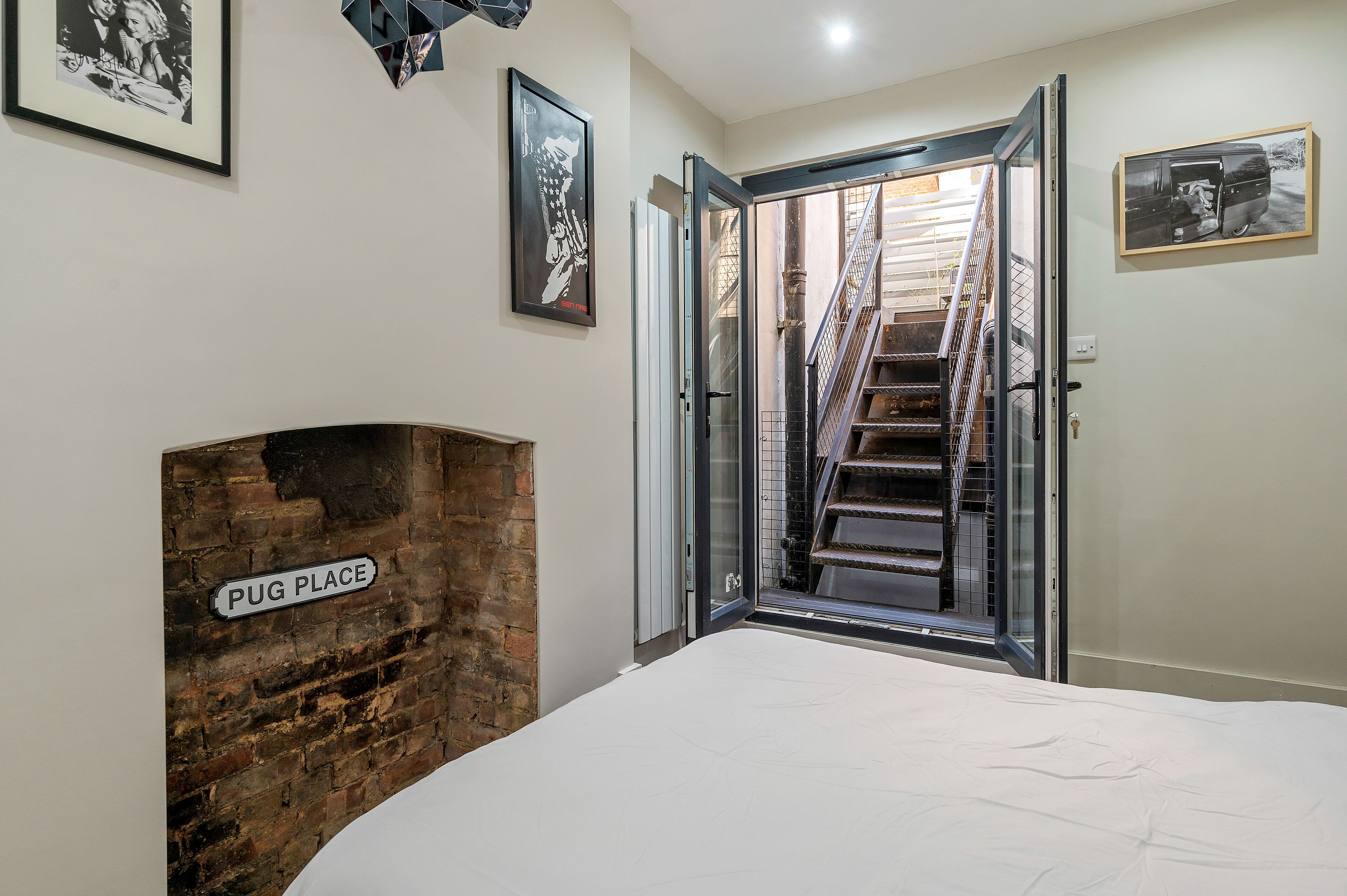 A bedroom with a brick fireplace labeled "PUG PLACE," a bed, a staircase leading to an outdoor area, and framed pictures on the walls.