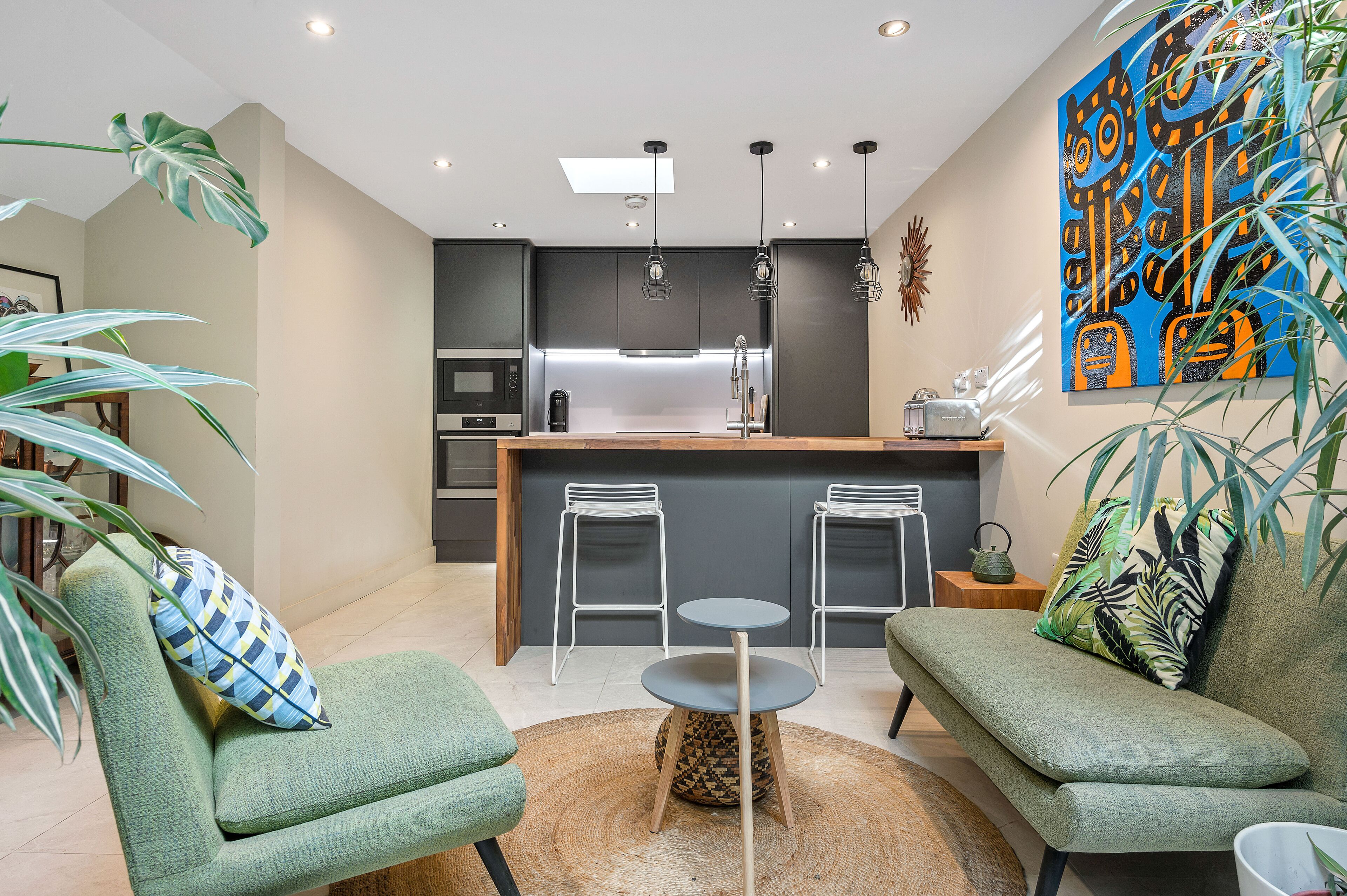 A modern living room with two green sofas, a small round table, and a kitchen area with a central island, bar stools, and a microwave.