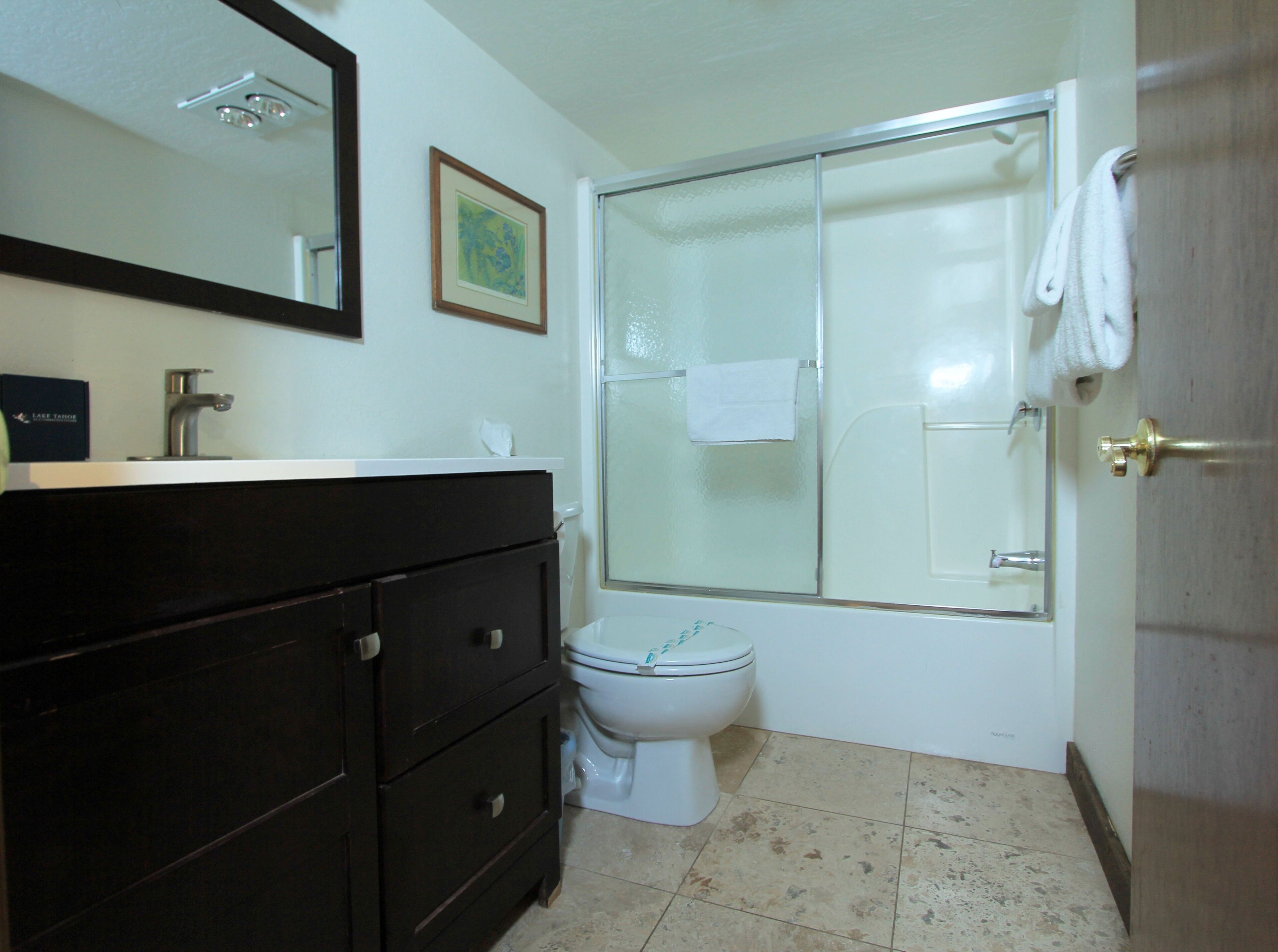 Un baño con tocador de madera oscura, inodoro blanco, cabina de ducha de vidrio y una toalla blanca colgada en la puerta de la ducha.